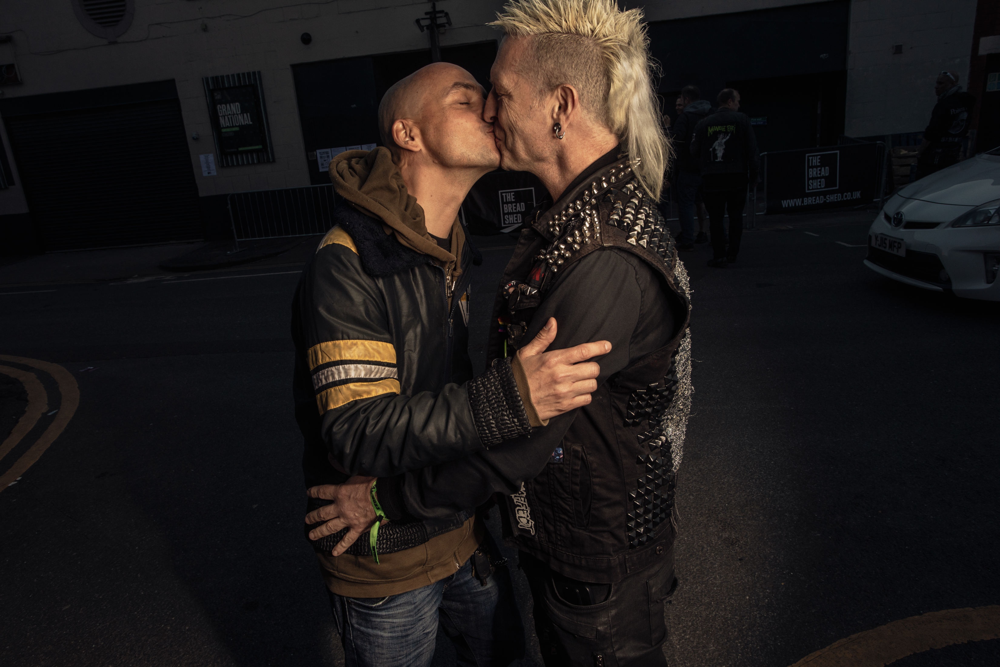 Two male punks kiss in a parking lot. The one on the left is wearing a leather jacket and hoodie, the one on the right has a studded leather vest and a mohawk.