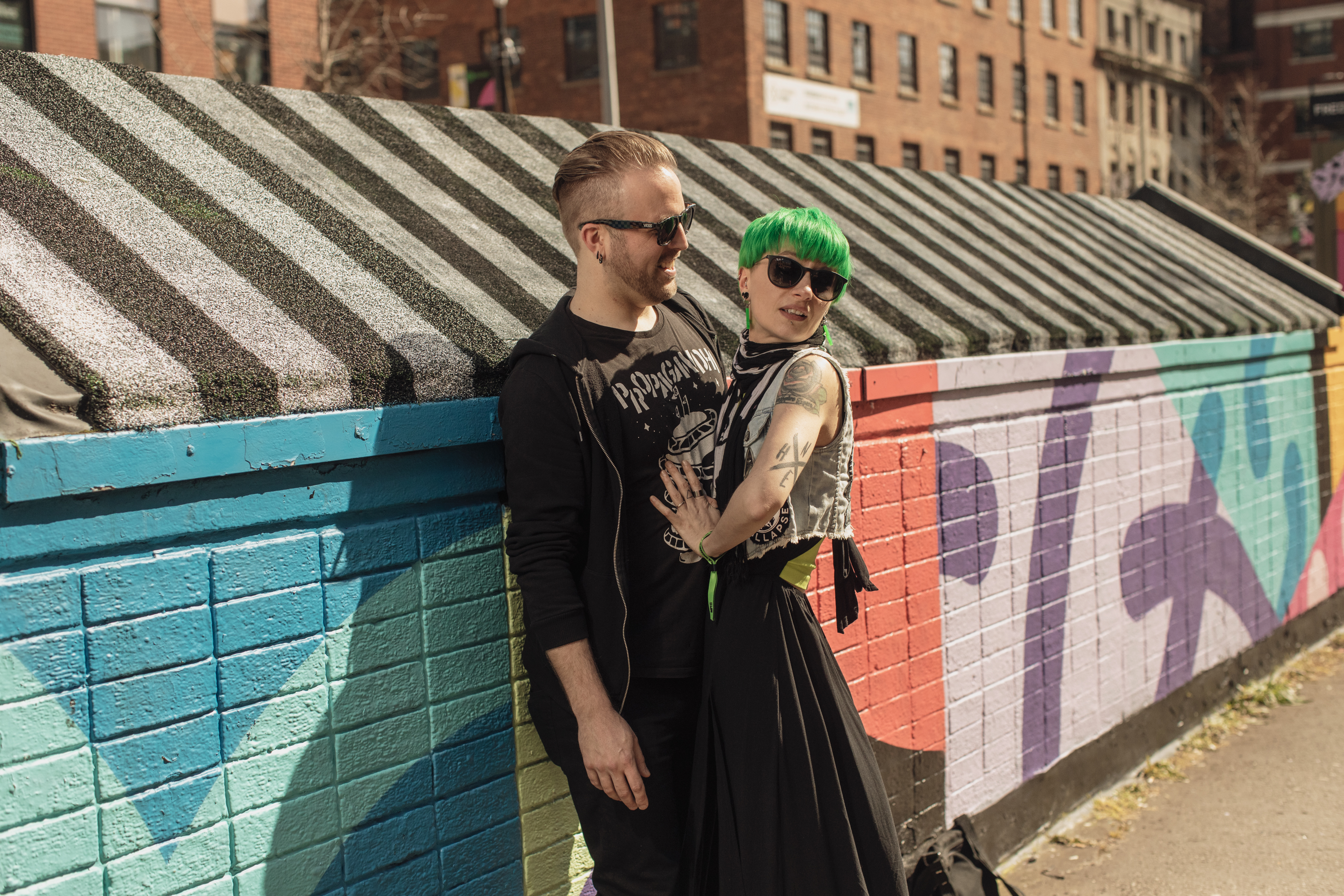 A guy wearing sunglasses and a band t-shirt is being embraced by a woman with bleached green hair and a denim vest. They're standing by a colorful mural on the street.