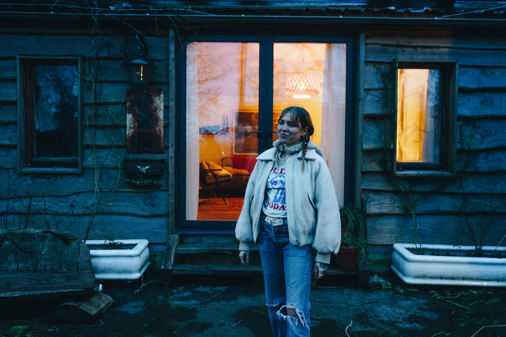 A young woman stands outside her self-built home at dusk, glass doors behind her.
