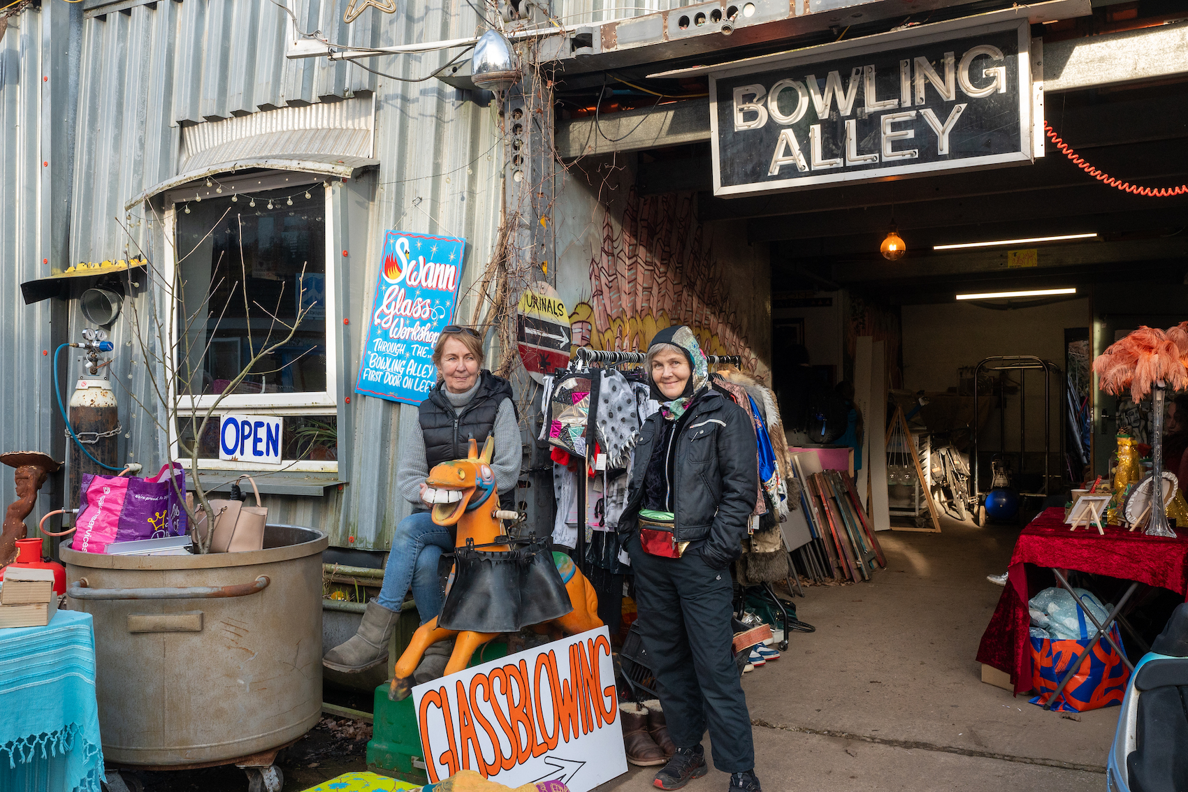Two people sit outside a shed surrounded by objects and a sign that says